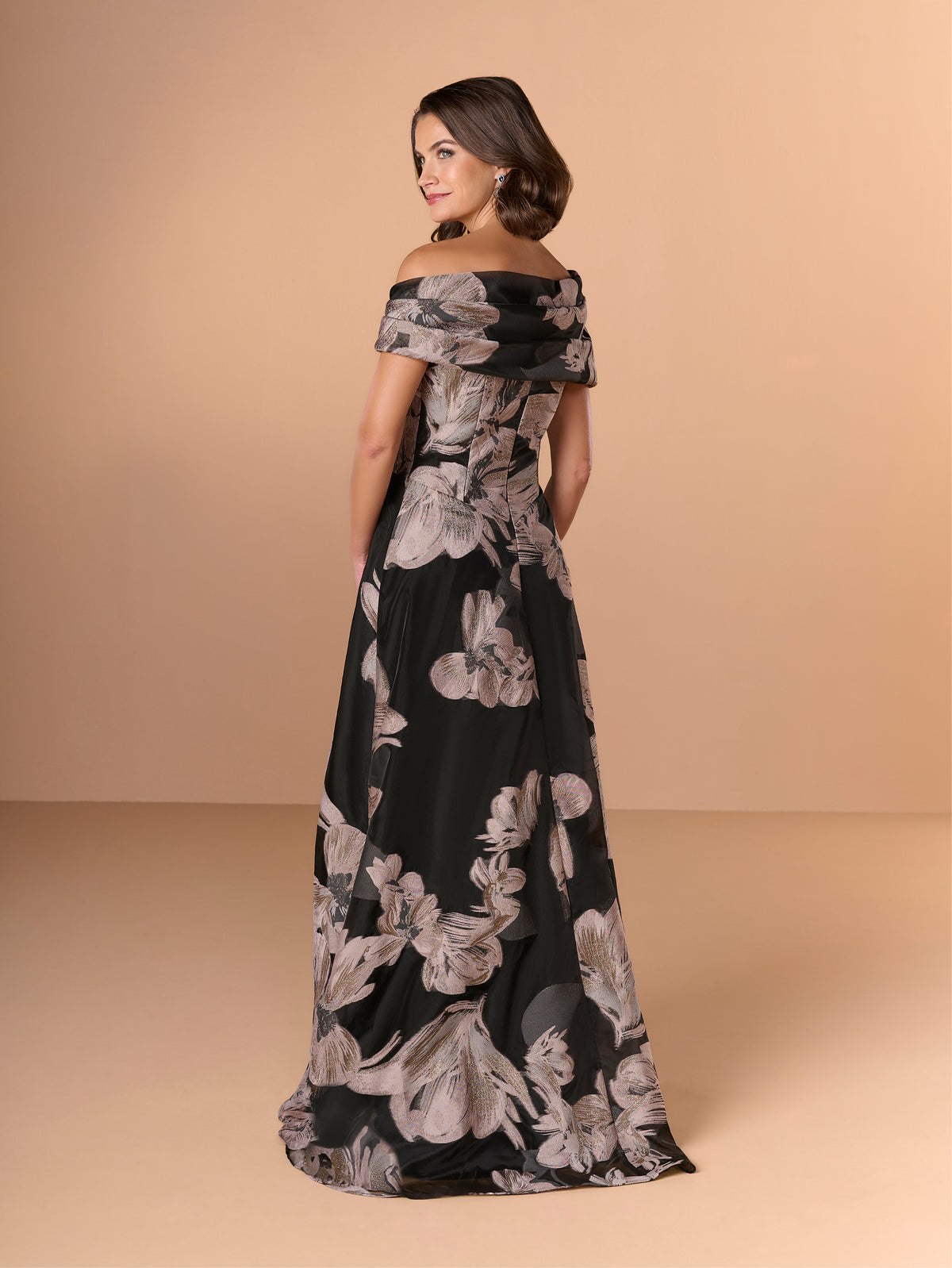 A woman modeling an off-the-shoulder floor-length gown with a black base and large gray floral print, photographed against a warm beige studio backdrop. / A-line style in textured organza with asymmetrical neckline and pleated wide collar off-shoulder.
