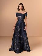 A woman modeling an off-the-shoulder navy blue floor-length gown with a shimmering floral pattern, set against a solid beige studio background. / A-line style in textured organza with asymmetrical neckline and pleated wide collar off-shoulder.