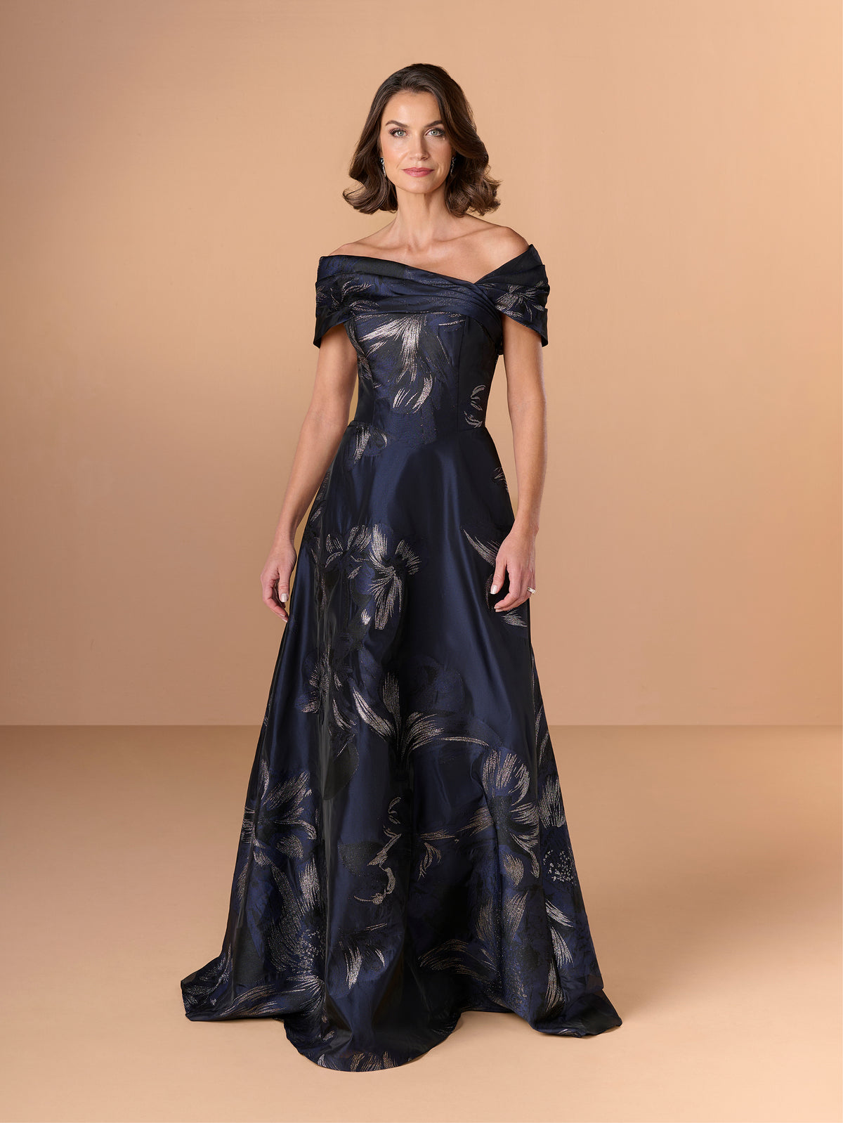 A woman modeling an off-the-shoulder navy blue floor-length gown with a shimmering floral pattern, set against a solid beige studio background. / A-line style in textured organza with asymmetrical neckline and pleated wide collar off-shoulder.