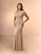 A woman modeling a floor-length champagne beige gown with intricate beaded floral embroidery and a V-neckline, set against a solid warm beige studio backdrop. / Slim long beaded style in soft mesh. V-neck and sheer short sleeves. Zipper back closure.