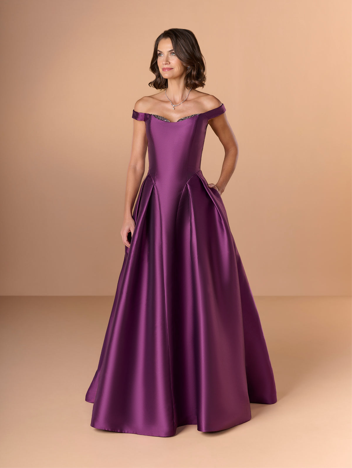 Woman wearing a purple off-shoulder gown against a beige background / Off the shoulder gown with side skirt panels and inserted neckline beading.
