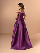 Woman wearing a purple off-shoulder gown against a beige background / Off the shoulder gown with side skirt panels and inserted neckline beading.