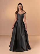 A woman modeling a floor-length off-the-shoulder ball gown in dark charcoal gray satin, set against a solid warm beige studio background. / Off the shoulder gown with side skirt panels and inserted neckline beading.
