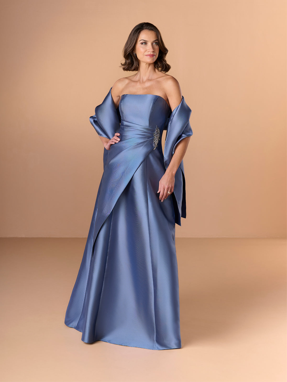 A woman modeling a floor-length satin gown in dusty blue. The off-the-shoulder dress features a structured bodice, draped sleeves, and an asymmetrical skirt, set against a solid warm beige studio background. / This strapless gown features a sleek, structured bodice that flows into an elegant A-line skirt. The design includes a dramatic, sculpted overlay at the waist, accented with a touch of sparkling embellishment on one side. Smooth, lustrous fabric and clean lines give the dress a sophisticated and moder
