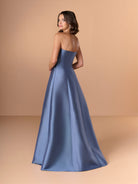 Woman wearing a strapless blue gown against a beige background / This strapless gown features a sleek, structured bodice that flows into an elegant A-line skirt. The design includes a dramatic, sculpted overlay at the waist, accented with a touch of sparkling embellishment on one side. Smooth, lustrous fabric and clean lines give the dress a sophisticated and modern look.