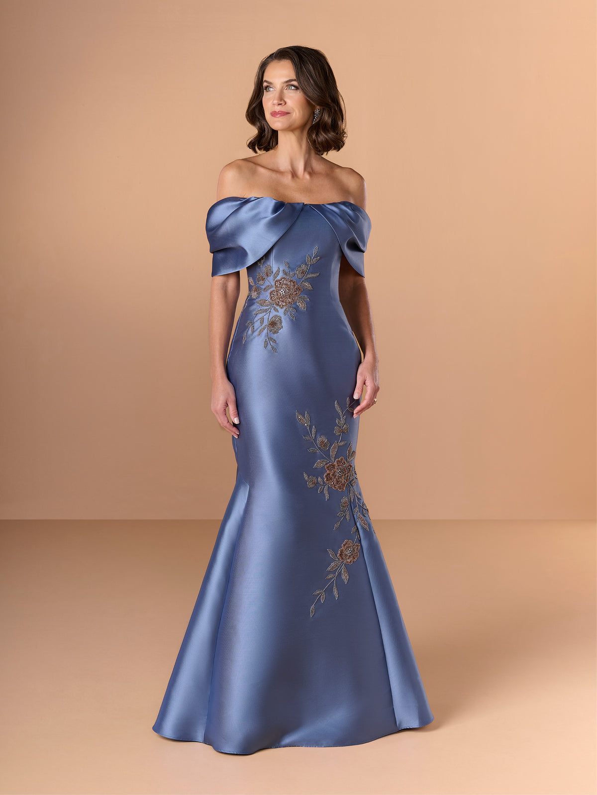 Woman wearing a blue off-shoulder gown with floral embroidery on a beige background / Off the shoulder mermaid gown with placed beading.