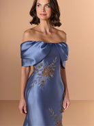 A woman wearing an off-the-shoulder slate blue satin dress with gold floral embroidery, posed against a solid beige background. / Off the shoulder mermaid gown with placed beading.