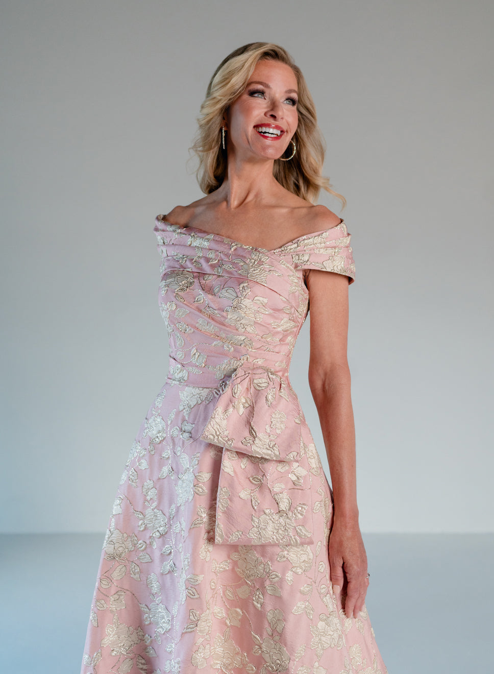 A woman modeling an off-the-shoulder light pink gown with intricate gold floral embroidery and a matching structured waist detail, photographed against a plain light gray studio background. / Brocade A-line style with off-shoulder collar/straps. Side front pleating on bodice finished with bow on side front waist area.
