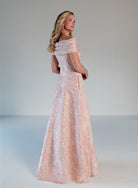 A woman modeling a floor-length off-the-shoulder gown in pale pink with a subtle iridescent floral pattern, photographed against a neutral light gray studio background. / Brocade A-line style with off-shoulder collar/straps. Side front pleating on bodice finished with bow on side front waist area.