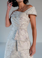 An off-the-shoulder champagne-colored dress with intricate floral embroidery and a structured bodice, shown on a model against a neutral studio background. / Brocade A-line style with off-shoulder collar/straps. Side front pleating on bodice finished with bow on side front waist area.