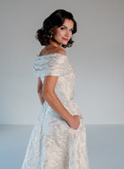 A woman modeling an off-the-shoulder, off-the-hip A-line gown in a textured cream/off-white fabric with a subtle floral jacquard pattern, set against a plain light gray studio background. / Brocade A-line style with off-shoulder collar/straps. Side front pleating on bodice finished with bow on side front waist area.