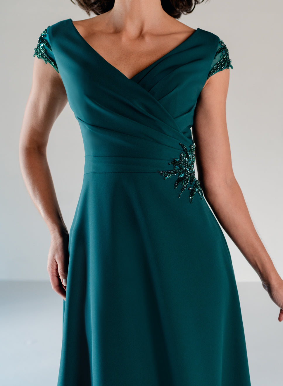 A woman wearing a teal green A-line dress with cap sleeves, embellished with beaded details on the shoulders and waist, photographed against a neutral light gray background. / A-line style in crepe with sheer beaded short cup sleeves. V-neck with side front pleating on bodice finished with beaded motif on side front waist area.