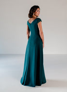 A woman modeling a floor-length teal evening gown from the back in a studio setting with a light gray background. / A-line style in crepe with sheer beaded short cup sleeves. V-neck with side front pleating on bodice finished with beaded motif on side front waist area.