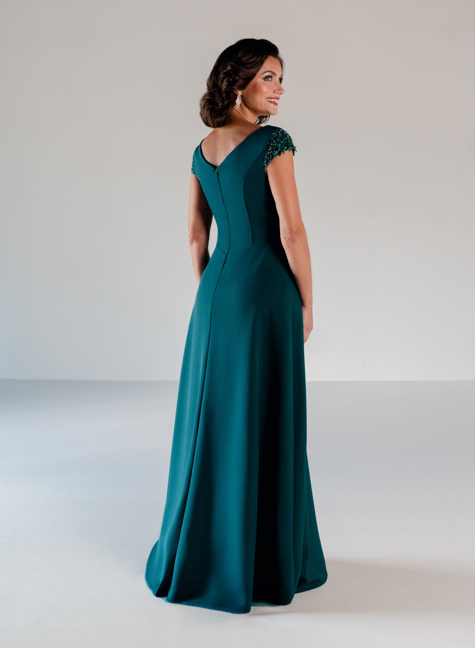 A woman modeling a floor-length teal evening gown from the back in a studio setting with a light gray background. / A-line style in crepe with sheer beaded short cup sleeves. V-neck with side front pleating on bodice finished with beaded motif on side front waist area.