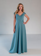 A woman modeling a floor-length teal blue gown with a draped V-neckline, cap sleeves, and lace applique at the waist, photographed against a neutral light gray studio background. / A-line style in crepe with sheer beaded short cup sleeves. V-neck with side front pleating on bodice finished with beaded motif on side front waist area.