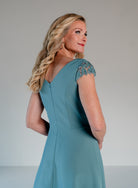 A woman modeling a sleeveless teal dress from the back, showing a V-neckline and lace-embellished cap sleeves, against a neutral studio background. / A-line style in crepe with sheer beaded short cup sleeves. V-neck with side front pleating on bodice finished with beaded motif on side front waist area.