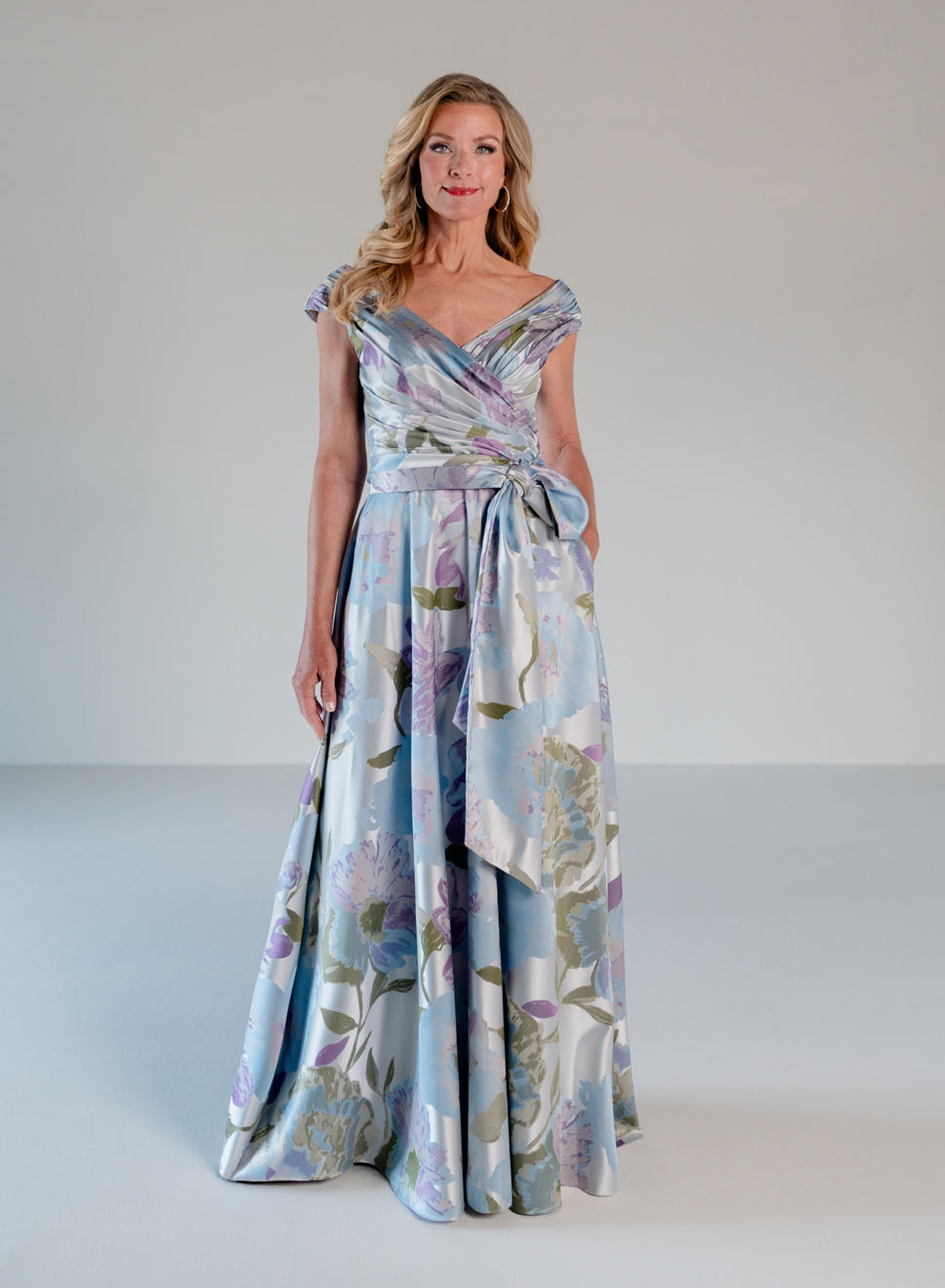 A woman modeling a floor-length light blue satin gown with a floral print in lavender, purple, and green, featuring a wrap-style bodice and a self-tie waist sash, set against a neutral studio background. / A-line style in jacquard. Pleated front bodice with V-neck and trim with bow on left side front waist area.