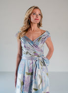 A woman modeling a light blue and white floral wrap dress with purple and green accents, set against a plain light gray studio background. / A-line style in jacquard. Pleated front bodice with V-neck and trim with bow on left side front waist area.