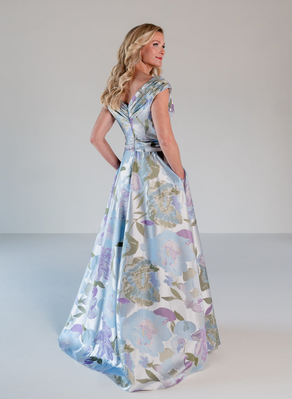 A woman modeling a floor-length satin gown with a floral print in light blue, lavender, and beige on a white base, photographed in a studio setting. / A-line style in jacquard. Pleated front bodice with V-neck and trim with bow on left side front waist area.