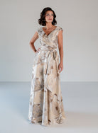 A woman modeling a floor-length wrap-style gown with a beige and taupe floral print, photographed against a plain light gray studio background. / A-line style in jacquard. Pleated front bodice with V-neck and trim with bow on left side front waist area.