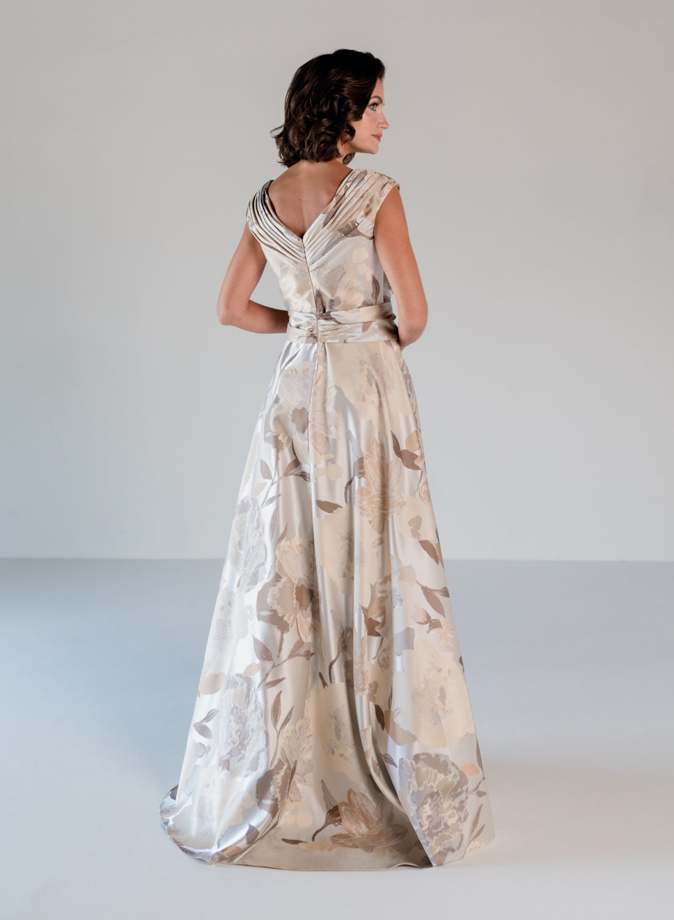 A woman wearing a floor-length formal gown with a draped, wrap-style back and a full skirt, shown from behind against a plain light gray studio background. / A-line style in jacquard. Pleated front bodice with V-neck and trim with bow on left side front waist area.