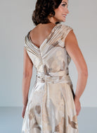 A woman wearing a sleeveless satin dress with a draped, pleated back and a matching waist tie, shown from the back in a studio setting. / A-line style in jacquard. Pleated front bodice with V-neck and trim with bow on left side front waist area.