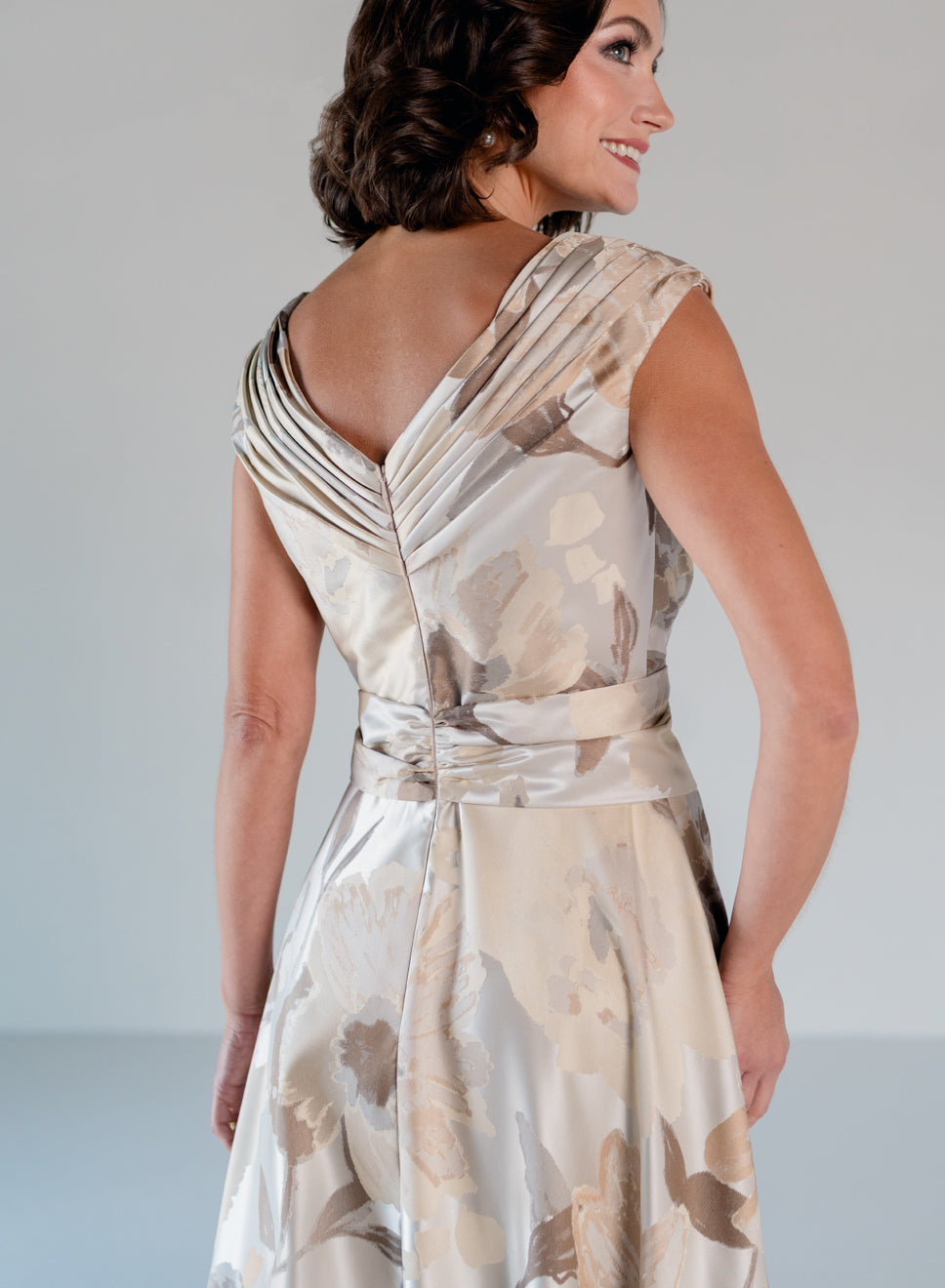 A woman wearing a sleeveless satin dress with a draped, pleated back and a matching waist tie, shown from the back in a studio setting. / A-line style in jacquard. Pleated front bodice with V-neck and trim with bow on left side front waist area.