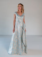 A woman modeling a floor-length formal gown with a shimmering light blue and gold floral pattern, standing against a neutral light gray studio background. / A-line style in soft brocade with V-neck and short cup sleeves, side draping on front bodice finished with draping panels on side front skirt.