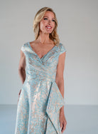 A woman modeling a light blue and silver patterned formal dress with a V-neckline and cap sleeves, set against a plain light gray studio background. / A-line style in soft brocade with V-neck and short cup sleeves, side draping on front bodice finished with draping panels on side front skirt.
