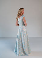 A woman modeling a floor-length light blue evening gown with a shimmering gold floral pattern, shown from the back in a studio setting with a neutral gray background. / A-line style in soft brocade with V-neck and short cup sleeves, side draping on front bodice finished with draping panels on side front skirt.