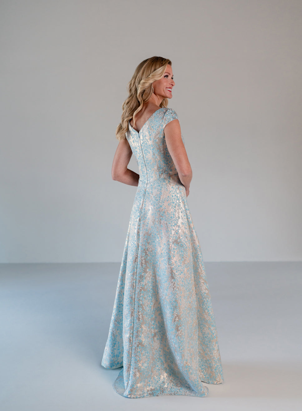 A woman modeling a floor-length light blue evening gown with a shimmering gold floral pattern, shown from the back in a studio setting with a neutral gray background. / A-line style in soft brocade with V-neck and short cup sleeves, side draping on front bodice finished with draping panels on side front skirt.