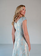 A woman modeling a light blue and silver floral lace dress, shown from a side-back angle against a neutral light gray studio background. / A-line style in soft brocade with V-neck and short cup sleeves, side draping on front bodice finished with draping panels on side front skirt.