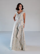 A woman modeling a floor-length formal gown with a shimmering silver and gold jacquard pattern, set against a plain light gray studio background. / A-line style in soft brocade with V-neck and short cup sleeves, side draping on front bodice finished with draping panels on side front skirt.