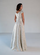 A woman wearing a floor-length champagne brocade evening gown, shown from the back in a studio setting with a light gray background. / A-line style in soft brocade with V-neck and short cup sleeves, side draping on front bodice finished with draping panels on side front skirt.