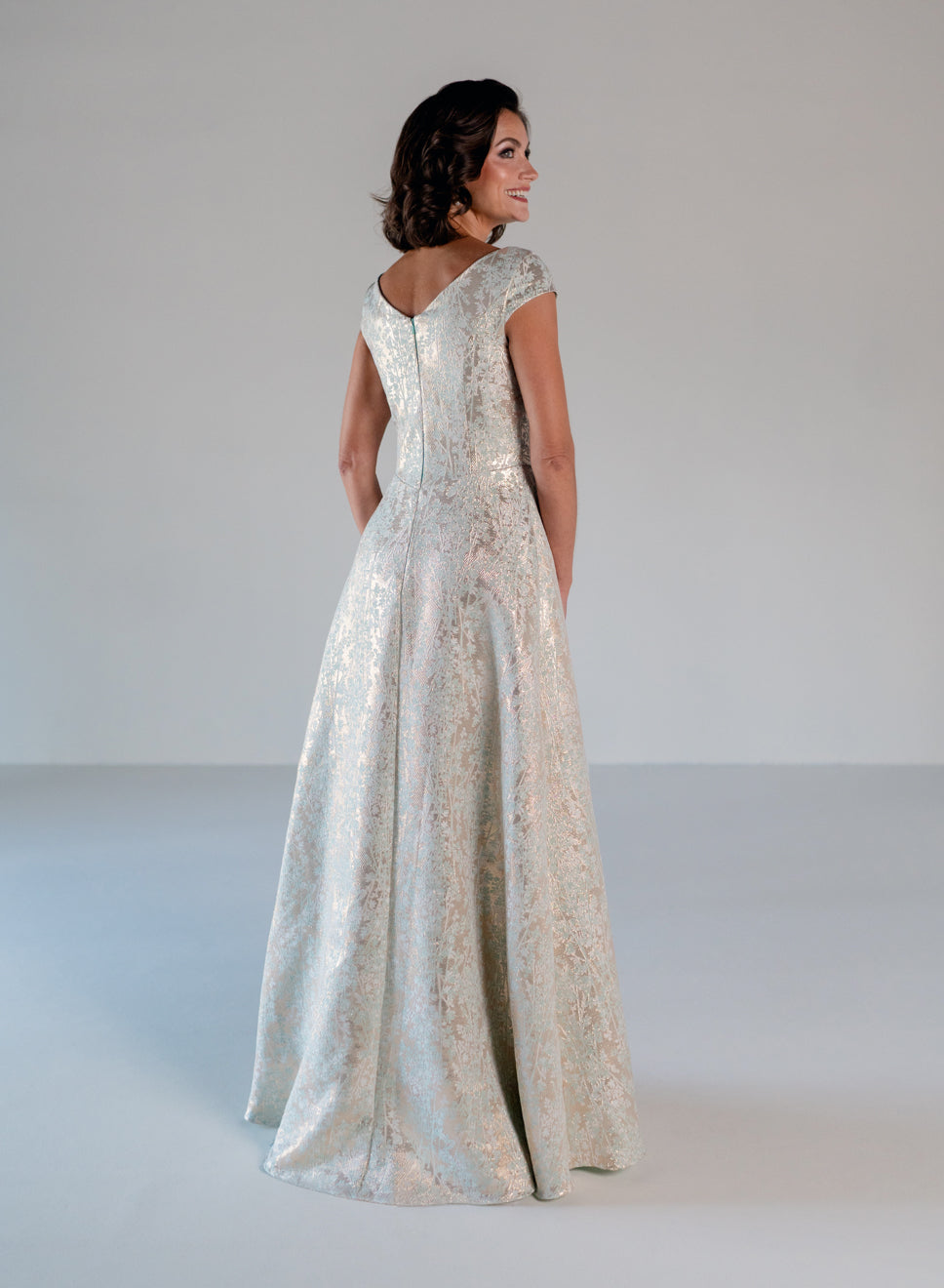 A woman wearing a floor-length champagne brocade evening gown, shown from the back in a studio setting with a light gray background. / A-line style in soft brocade with V-neck and short cup sleeves, side draping on front bodice finished with draping panels on side front skirt.