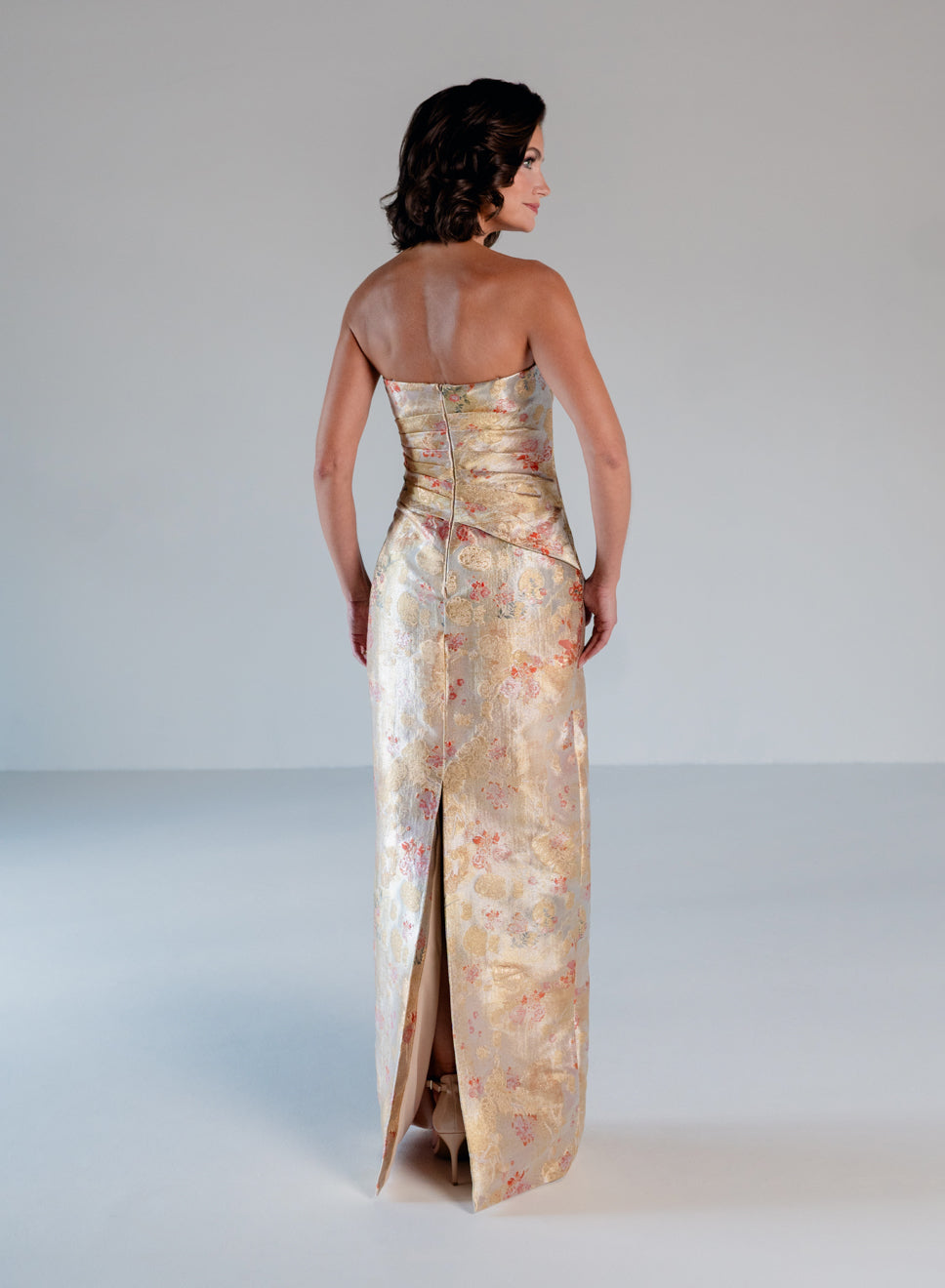 A woman stands with her back to the camera wearing a strapless floor-length gown with a fitted corset-style waist and a long slit. The dress has a shimmering champagne-gold base with a soft floral print in muted pink, peach, and green. / Jacquard style with straight neckline and side draping on front bodice. Pencil skirt with split on CB.