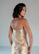 A woman shown from the back wearing a strapless champagne-gold satin dress with a subtle floral and circular jacquard pattern, set against a plain light gray studio background. / Jacquard style with straight neckline and side draping on front bodice. Pencil skirt with split on CB.