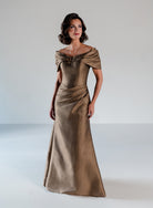 A woman modeling a floor-length off-the-shoulder gown in a shimmering metallic taupe-gold fabric against a plain light gray studio background. / A-line style in shimmer jacquard with off-shoulder collar and side front draping on skirt.