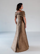 A woman modeling a floor-length metallic bronze gown with a draped off-the-shoulder neckline, shown from the back in a studio setting. / A-line style in shimmer jacquard with off-shoulder collar and side front draping on skirt.