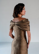 A woman shown from the back wearing a metallic champagne-gold off-the-shoulder evening gown with a structured corset back and draped fabric detail. / A-line style in shimmer jacquard with off-shoulder collar and side front draping on skirt.