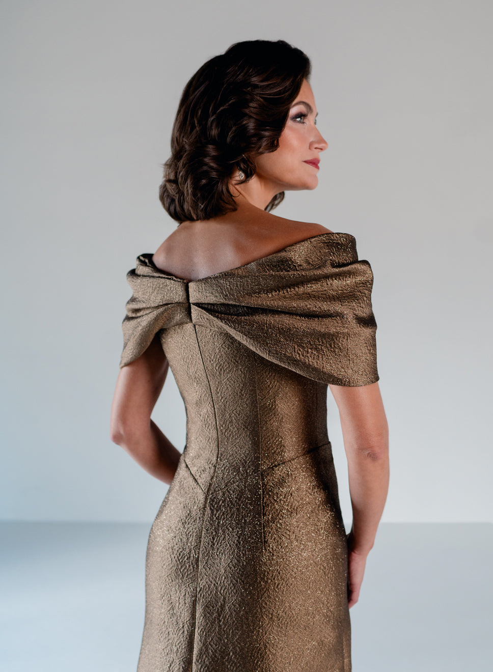 A woman shown from the back wearing a metallic champagne-gold off-the-shoulder evening gown with a structured corset back and draped fabric detail. / A-line style in shimmer jacquard with off-shoulder collar and side front draping on skirt.