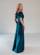 A woman modeling a floor-length off-the-shoulder gown in a rich teal emerald green fabric, photographed against a neutral light gray studio background. / A-line style in shimmer jacquard with off-shoulder collar and side front draping on skirt.