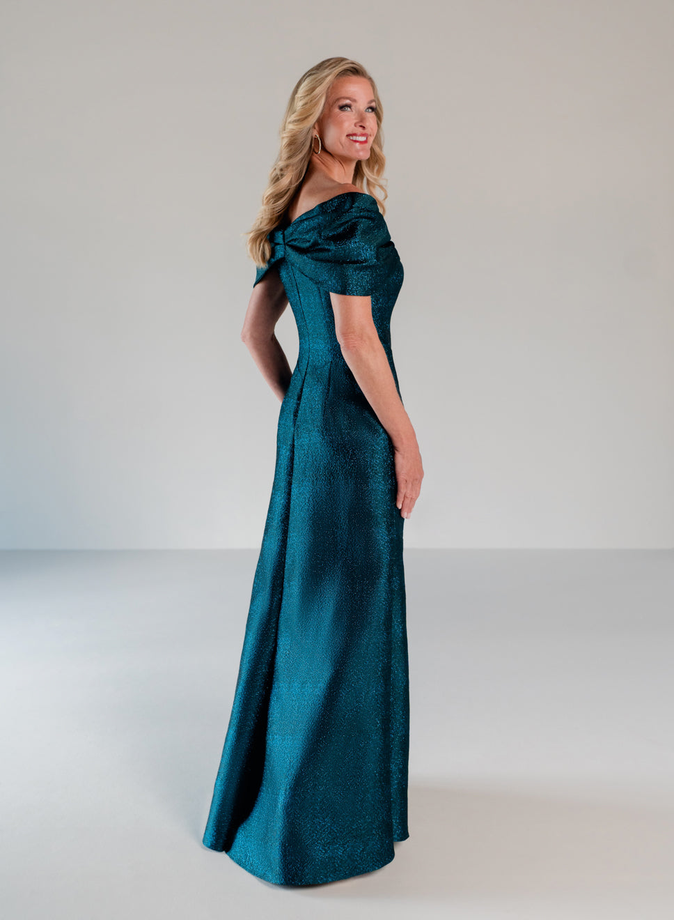A woman modeling a floor-length off-the-shoulder gown in a rich teal emerald green fabric, photographed against a neutral light gray studio background. / A-line style in shimmer jacquard with off-shoulder collar and side front draping on skirt.