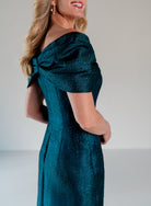 A woman wearing a shimmering teal off-the-shoulder dress with short sleeves and a structured bow detail at the neckline, shown from a side-back angle against a plain light gray studio background. / A-line style in shimmer jacquard with off-shoulder collar and side front draping on skirt.