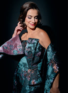 A woman wearing a strapless off-the-shoulder gown with a teal floral jacquard pattern and a matching draped capelet, posed against a dark studio background. / Slim style in jacquard with straight neckline and large bow on side front decorated with small, beaded detail.