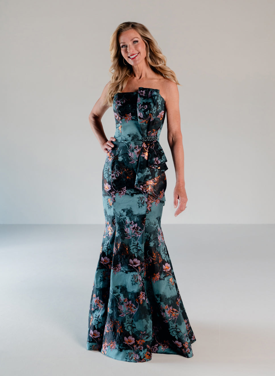 A woman modeling a strapless mermaid-style gown with a floral print in teal, black, and coral tones against a plain light gray studio background. / Slim style in jacquard with straight neckline and large bow on side front decorated with small, beaded detail.