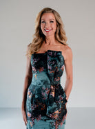 A smiling blonde woman wearing a strapless fitted dress with a dark teal and black floral print, ruffled trim, and a jeweled waist detail, photographed against a light gray studio background. / Slim style in jacquard with straight neckline and large bow on side front decorated with small, beaded detail.