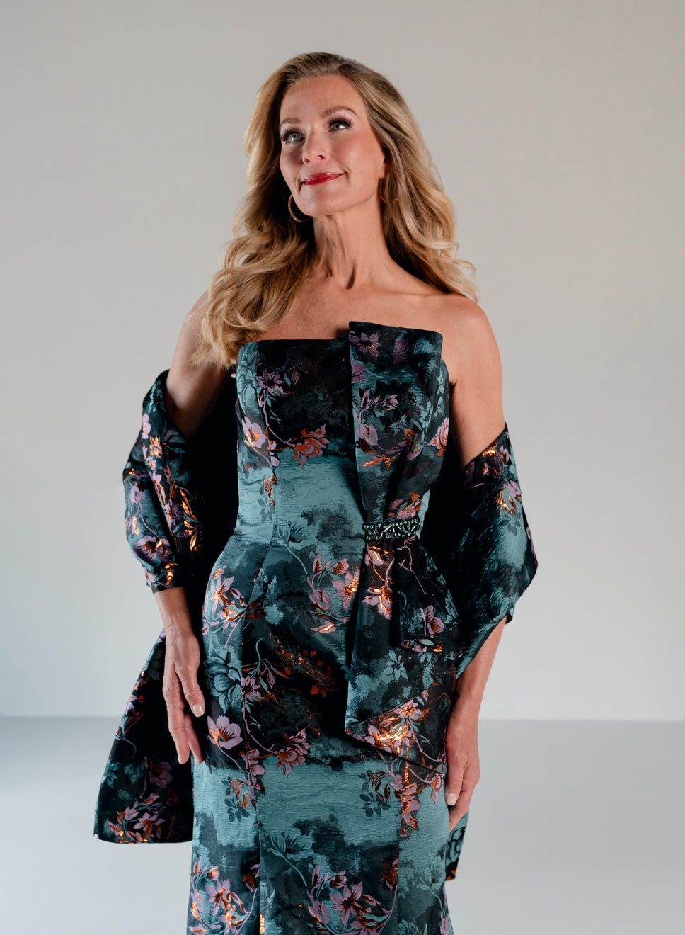 A woman modeling an off-the-shoulder floral dress with a structured bodice and long sleeves, photographed against a plain light gray studio background. / Slim style in jacquard with straight neckline and large bow on side front decorated with small, beaded detail.