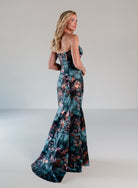 A woman modeling a strapless mermaid gown with a dark teal floral print, photographed in a studio setting against a light gray background. / Slim style in jacquard with straight neckline and large bow on side front decorated with small, beaded detail.