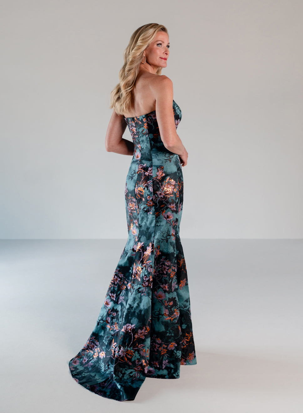 A woman modeling a strapless mermaid gown with a dark teal floral print, photographed in a studio setting against a light gray background. / Slim style in jacquard with straight neckline and large bow on side front decorated with small, beaded detail.
